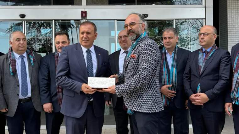 Bir grup erkek, bir binanın önünde duruyor; biri diğerine bir plaket veriyor.