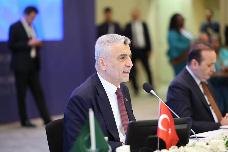 Toplantı masasında mikrofonla konuşan takım elbiseli bir kişi, önünde Türk ve yeşil bayraklarla görülüyor.