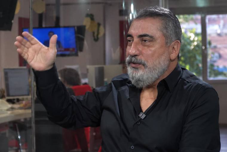 İç mekânda oturan sakallı bir kişi, bir elini kaldırarak konuşuyor; arka planda ekran ve çalışma masaları var.