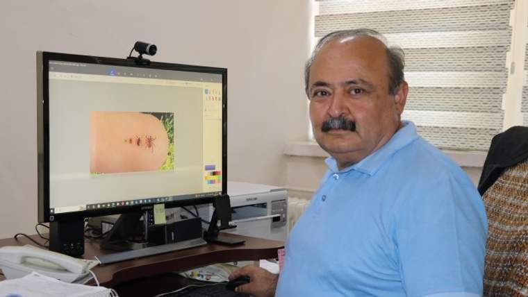 Bilgisayar ekranında kene görüntüsü açıkken masada oturan bıyıklı bir kişi ve ofis ekipmanları görülüyor.