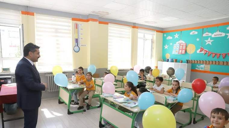Sınıfta takım elbiseli bir kişi, renkli balonlarla süslenmiş sıralarda oturan öğrencilerin yanında duruyor.