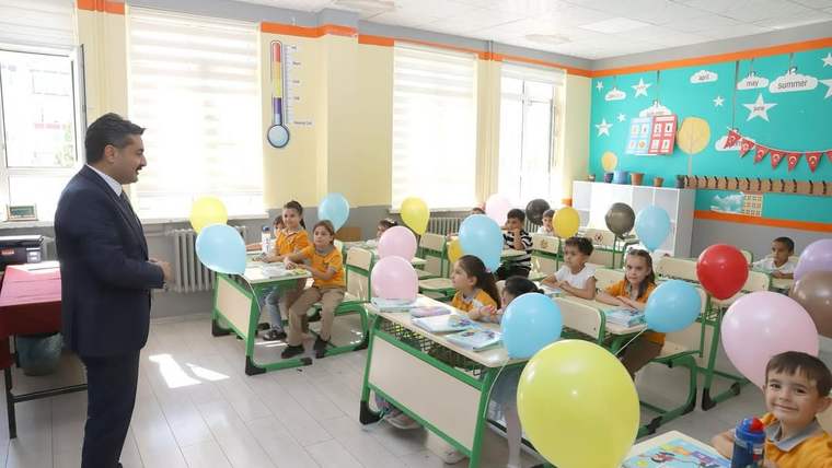Sınıfta takım elbiseli bir kişi, renkli balonlarla süslenmiş sıralarda oturan öğrencilerin yanında duruyor.