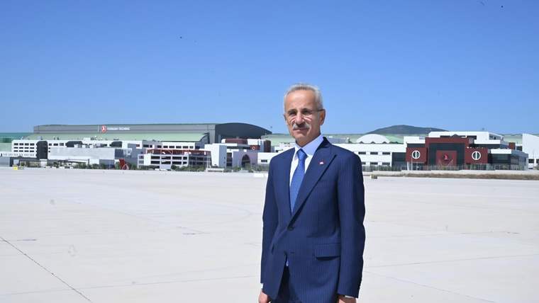 Takım elbiseli bir kişi, havaalanı apronunda; arka planda terminal binaları ve dağlık arazi görülüyor.