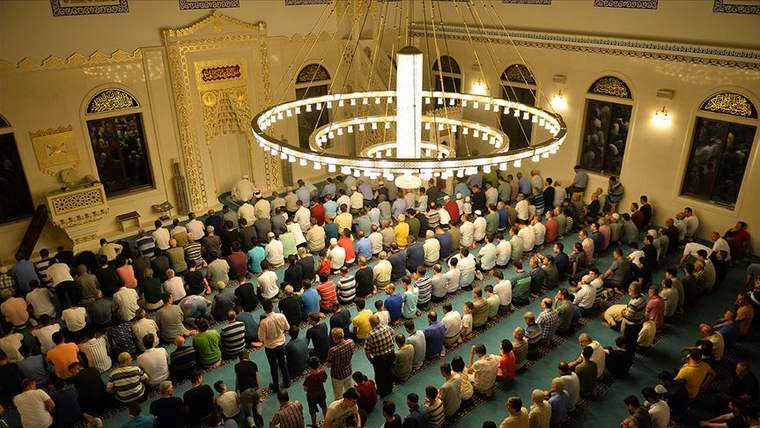 Cami içinde teravih namazı kılan kalabalık bir cemaat, avizelerle aydınlatılmış.