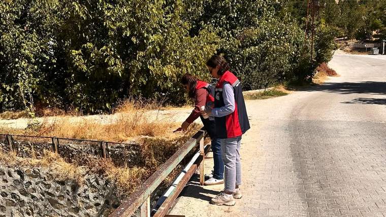 İki kişi, su kaynağının yanında inceleme yapıyor. Etraf yeşil bitki örtüsüyle kaplı.