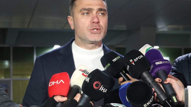 Koyu ceketli bir kişi, bina girişinde DHA ve İHA logolu mikrofonlara konuşuyor.