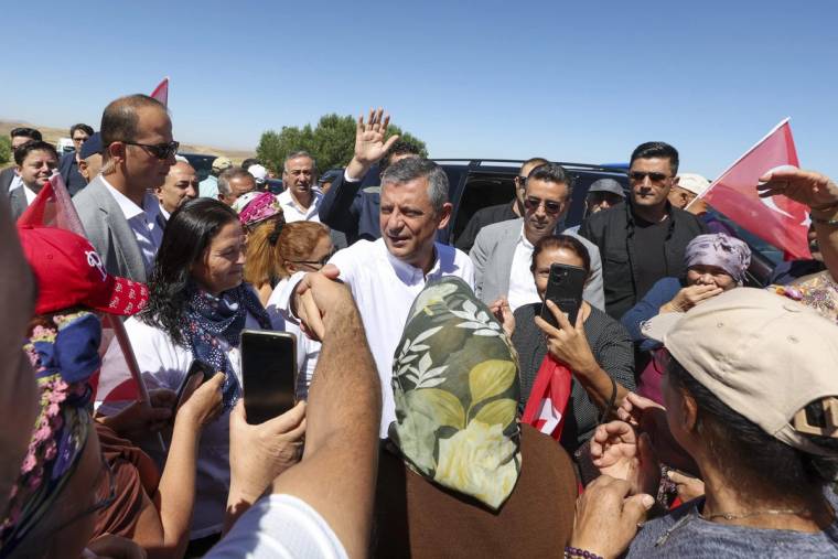 Açık havada, beyaz gömlekli bir kişi çevresindeki kalabalık ve telefonlarla görüntülenirken kırmızı bayraklar görülüyor.