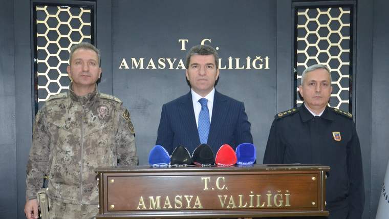 kürsüde mikrofonlar önünde duran takım elbiseli kişi, iki üniformalı kişinin arasında görülüyor.