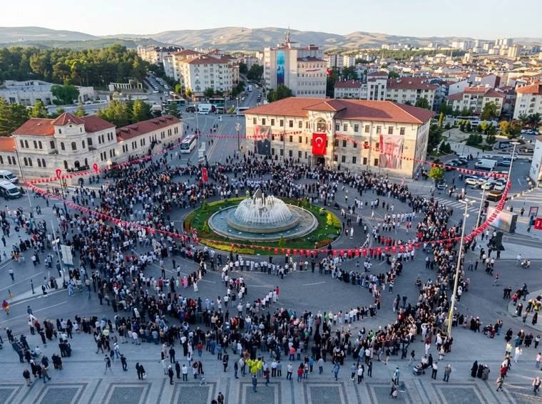Havadan görülen geniş meydanda, çeşme çevresinde toplanan kalabalık ve kırmızı-beyaz süslemeler yer alıyor.