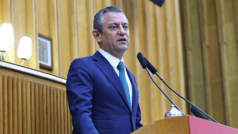 Ahşap panelli bir salonda takım elbiseli bir kişi, kürsüde iki mikrofonun önünde konuşuyor.