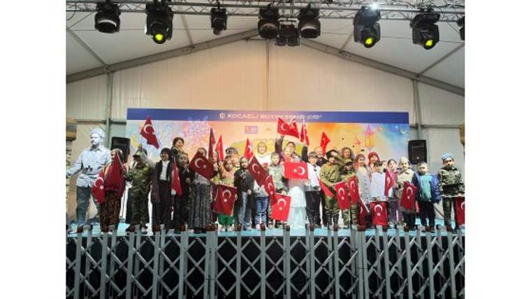Sahnede çocuk ve yetişkinlerden oluşan grup, ellerinde Türk bayrakları ve ay yıldızlı flamalarla duruyor.