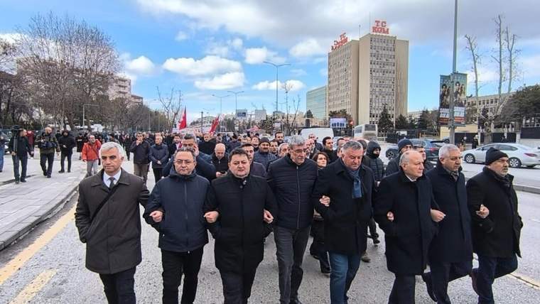 Kollarını birbirine kenetleyen bir grup erkek, arkalarındaki kalabalık ve bayraklarla kent caddesinde yürüyor.