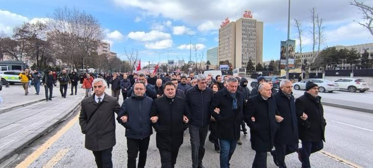 Kollarını birbirine kenetleyen bir grup erkek, arkalarındaki kalabalık ve bayraklarla kent caddesinde yürüyor.