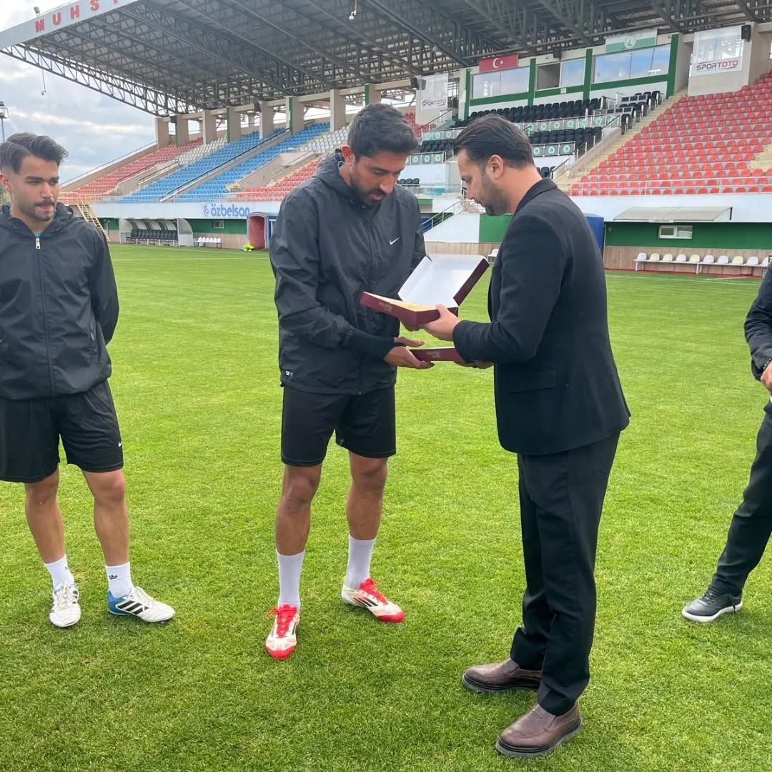 Futbol sahasında iki kişi, diğer takım üyelerinin yanında açık bir dosya ve kitap inceliyor.