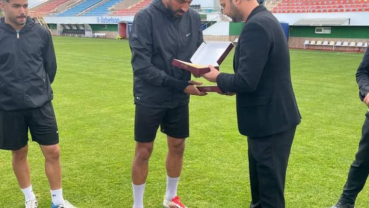 Futbol sahasında iki kişi, diğer takım üyelerinin yanında açık bir dosya ve kitap inceliyor.