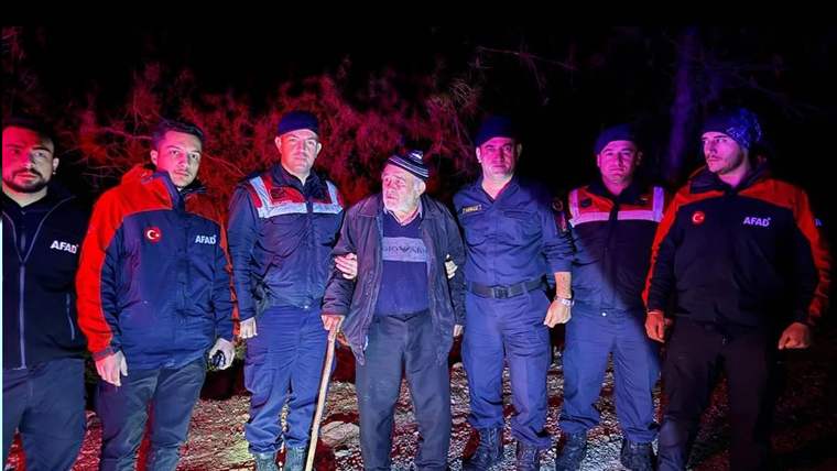 Gece karanlığında AFAD yazılı üniformalı görevliler, yaşlı bir adamla birlikte ormanlık alanda duruyor.