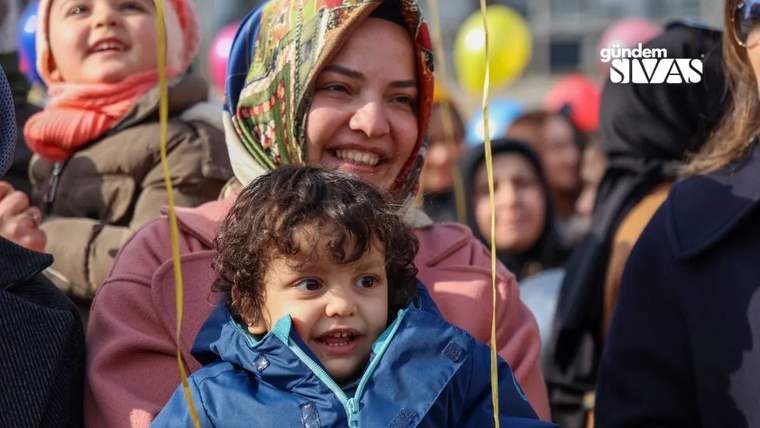 Gülümseyen bir kadın, mavi montlu küçük bir çocuğu kucağında tutuyor; arkada kalabalık ve balonlar görülüyor.
