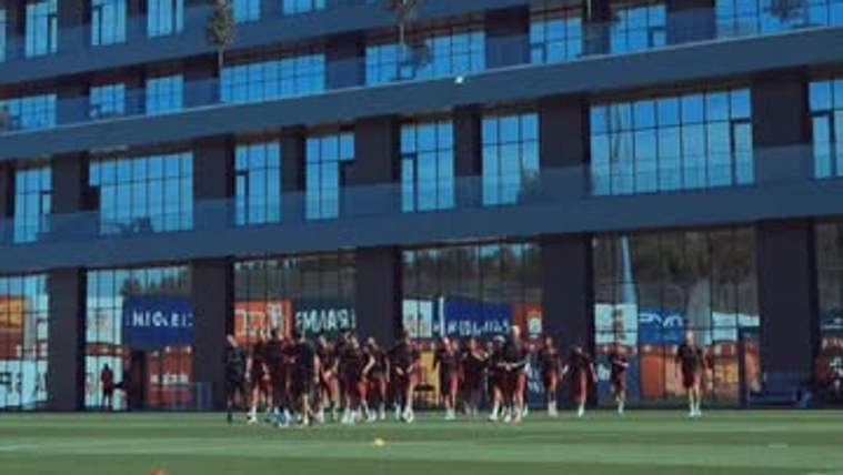 Cam cepheli çok katlı bir binanın önünde, futbol sahasında kırmızı formalı bir grup oyuncu duruyor.