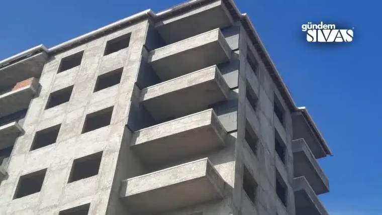 Tamamlanmamış bir apartman binası, 9. katı görünür şekilde. Bina gri renkte ve pencereleri açık.