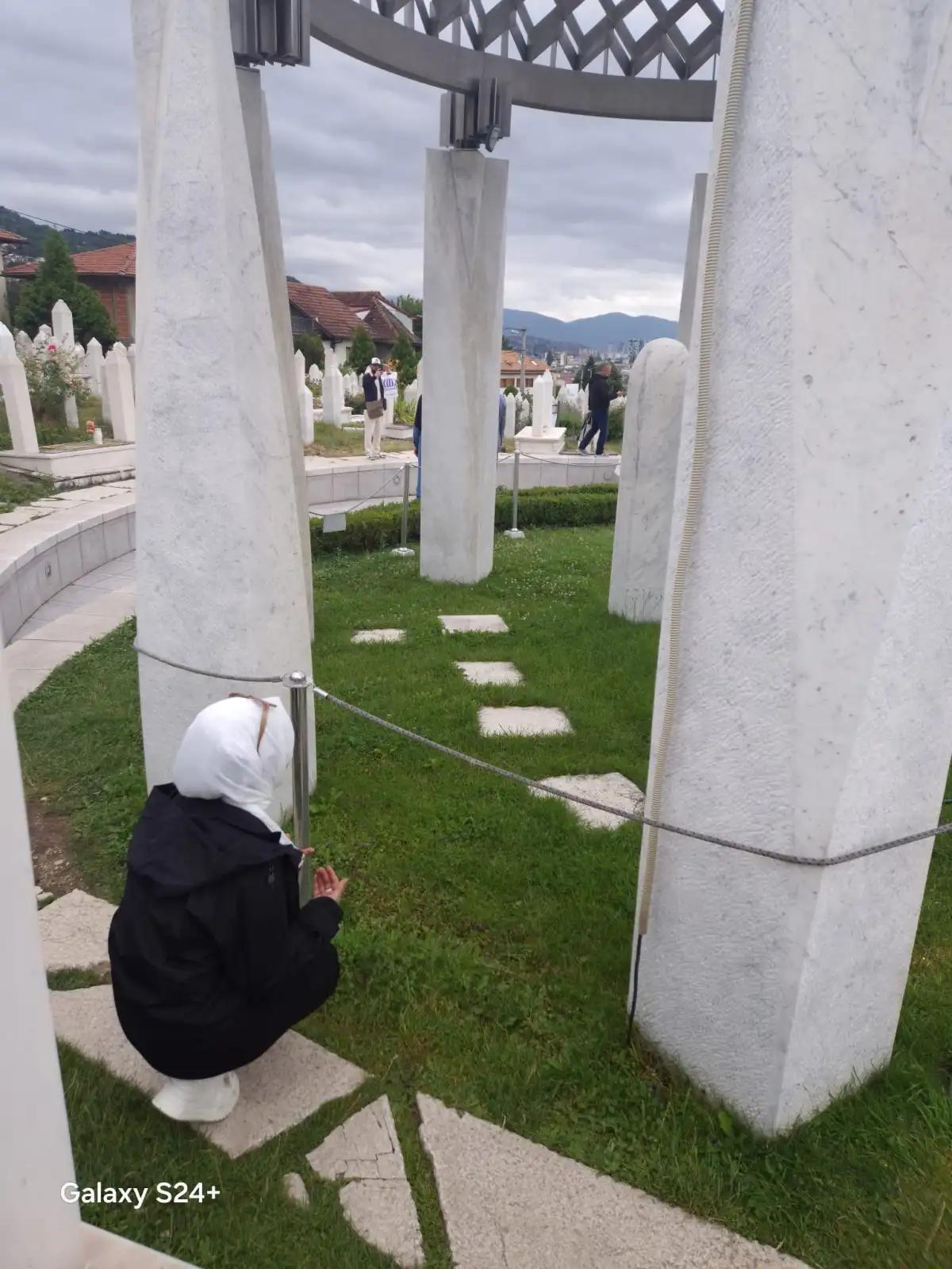Beyaz taşlardan oluşan mezarların bulunduğu bir mezarlıkta, başı örtülü bir kadın dua ediyor.