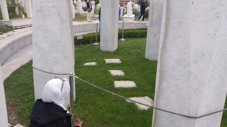 Beyaz taşlardan oluşan mezarların bulunduğu bir mezarlıkta, başı örtülü bir kadın dua ediyor.