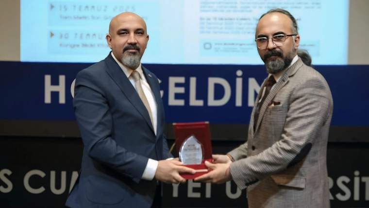 Sahnede iki erkek, üzerinde plaket bulunan kırmızı kutuyu birlikte tutuyor; arkalarında sunum perdesi görülüyor.