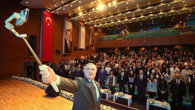 Bir kişi, selfie çubuğu ile fotoğraf çekiyor; arka planda kalabalık bir izleyici ve Türk bayrağı görünmekte.