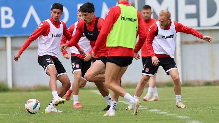 Yeşil sahada kırmızı formalar ve beyaz antrenman yelekleri giyen futbolcular topun çevresinde koşuyor.