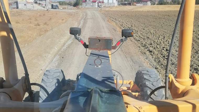 Bir iş makinesi, stabilize yolun üzerinde ilerliyor; yolun iki yanında tarla ve yapılar görünmekte.