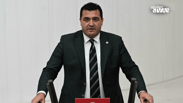 Kürsüde konuşan bir kişi, siyah takım elbise giymekte ve mikrofonun önünde durmakta.