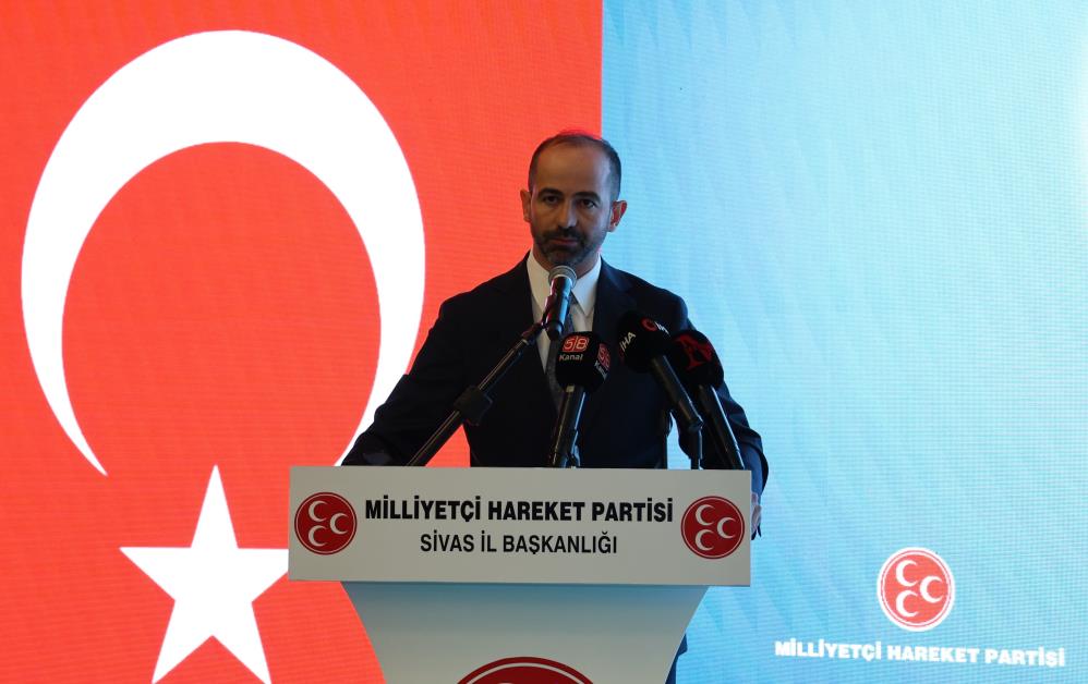 Kürsüde konuşan bir kişi, üzerinde Milliyetçi Hareket Partisi Sivas İl Başkanlığı yazan pankartın önünde duruyor.