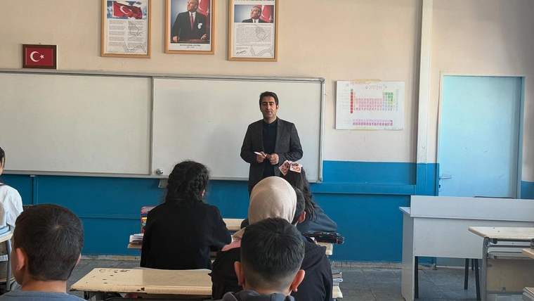 Sınıfta bir kişi, sıralarında oturan öğrenci grubunun karşısında duruyor; duvarda Türk bayrağı ve portreler bulunuyor.