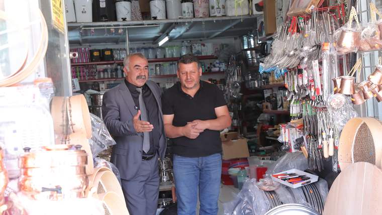 Hırdavat dükkânında iki erkek, raflardaki çeşitli ürünlerin önünde ayakta duruyor; biri el hareketi yapıyor.