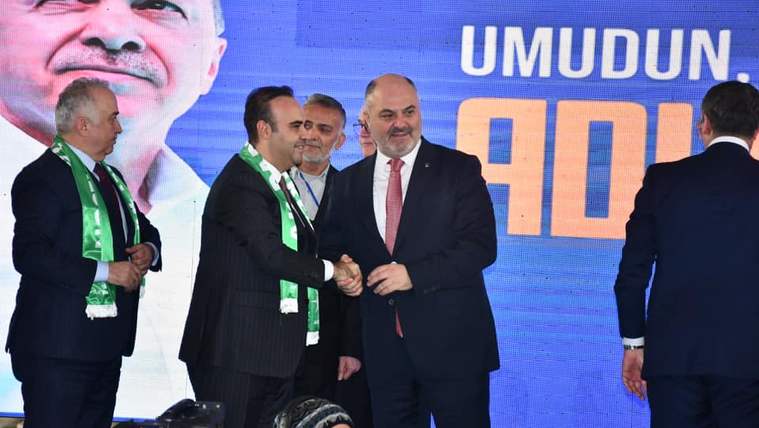 Sahnede dört erkekten ikisi el sıkışırken arkadaki ekranda büyük bir portre ve “UMUDUN” yazısı görülüyor.