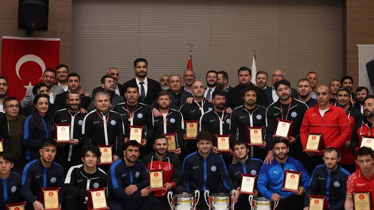 Bir salonda çok sayıda kişi, ellerinde plaketlerle ve önlerinde kupalarla birlikte poz veriyor.