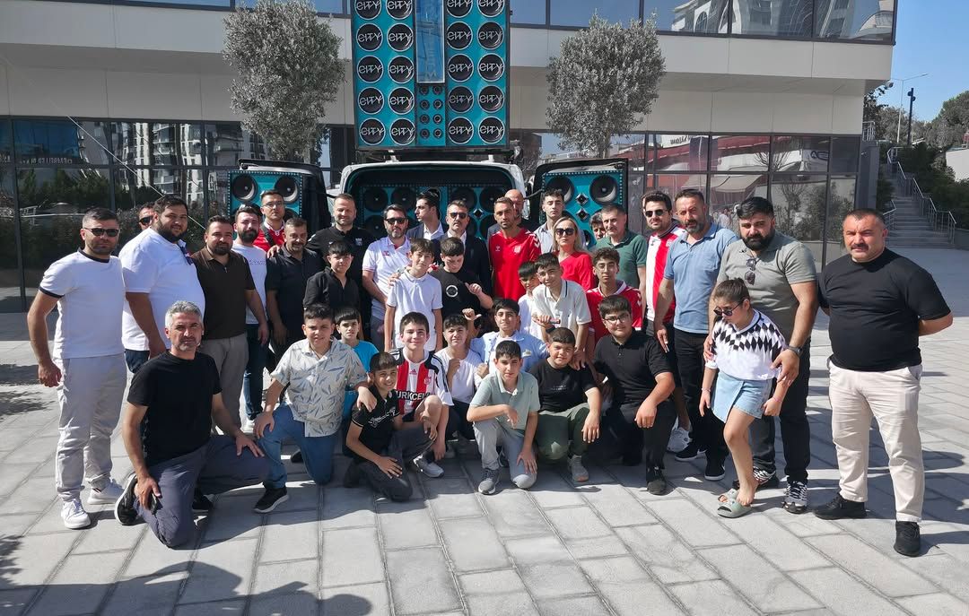 Bir bina önünde, aralarında kırmızı-beyaz formalar giyen çocukların da bulunduğu kalabalık grup poz veriyor.