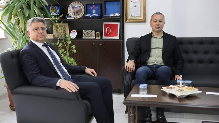 İki kişi, bir ofiste oturuyor; arka planda çeşitli ödüller ve belgeler görünmekte.