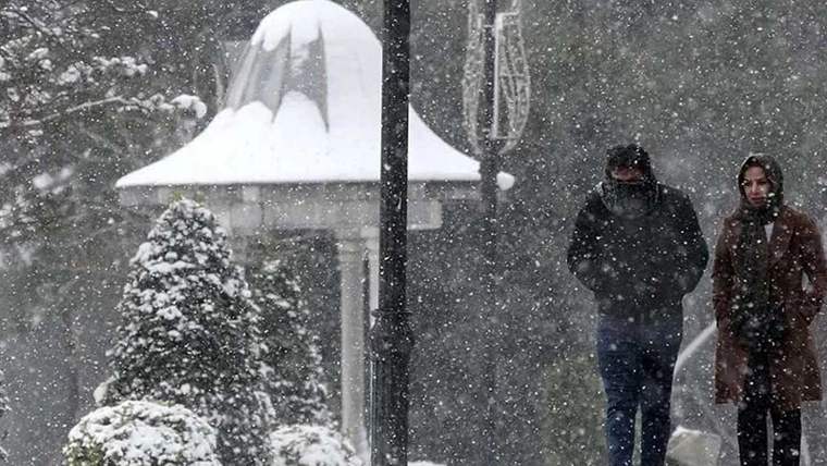 Yoğun kar yağışı altında kapüşonlu iki kişi yürürken, karla kaplı ağaç ve beyaz çatılı yapı görülüyor.