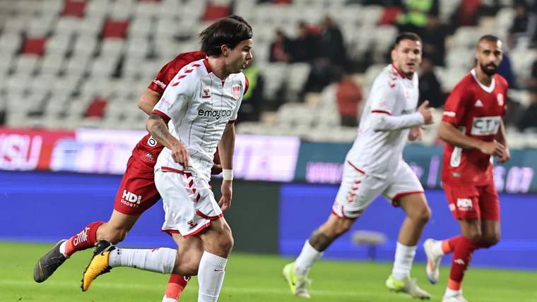 Beyaz forma giymiş bir futbolcu, topu sürerek ilerliyor; arka planda diğer futbolcular yer alıyor.