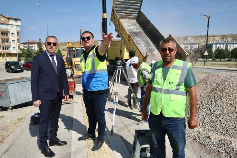 İki kişi, inşaat alanında çalışırken bir kişi yön gösteriyor. Arkada iş makineleri ve malzeme yığını var.