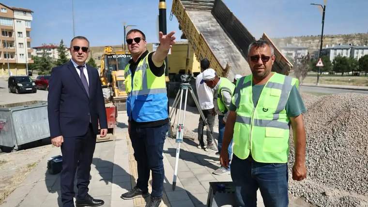 İki kişi, inşaat alanında çalışırken bir kişi yön gösteriyor. Arkada iş makineleri ve malzeme yığını var.