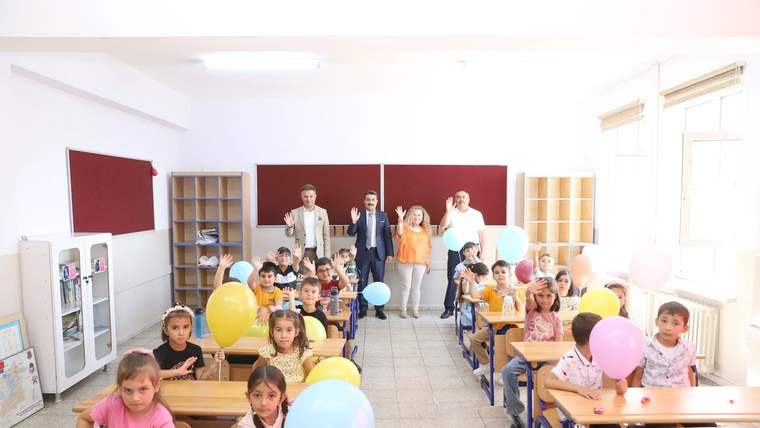 Sınıfta sıralarında oturan çocuklar ellerinde renkli balonlar tutarken arka planda yetişkinler duruyor.