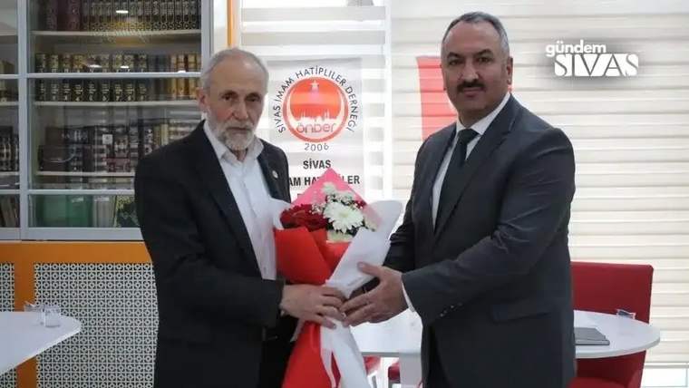 İki erkek, takım elbiseleriyle bir buketi birlikte tutarken arka planda dernek logosu görülüyor.