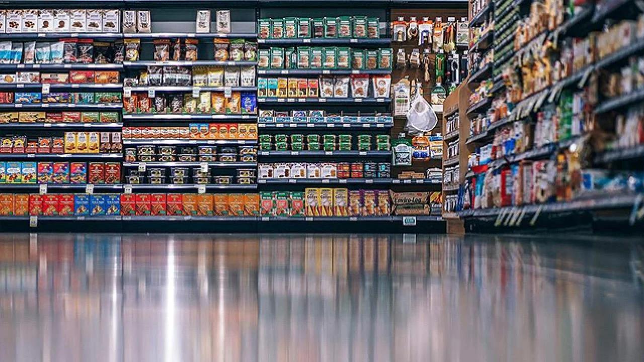 Market koridorunda çeşitli ambalajlı gıdalarla dolu raflar ve zeminde ışık yansımaları görülüyor.