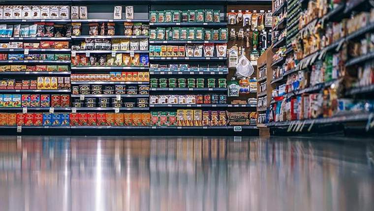 Market koridorunda çeşitli ambalajlı gıdalarla dolu raflar ve zeminde ışık yansımaları görülüyor.
