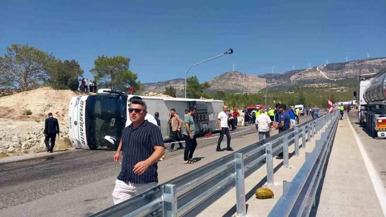 Yol kenarında yan yatmış bir otobüs, çevresinde yürüyen insanlar ve korkuluklar görülüyor; arkada dağlar uzanıyor.