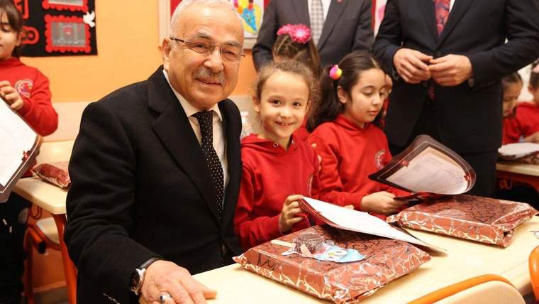 Takım elbiseli bir kişi, sınıfta kırmızı giysili çocuklarla birlikte masada paketler ve belgelerle görülüyor.