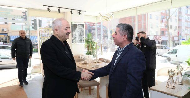 Aydınlık bir mekânda iki erkek el sıkışırken, arkada üç kişi ve masa düzeni görülüyor.