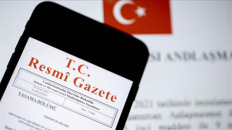 Bir cep telefonunun ekranında T.C. Resmi Gazete belgesi görünüyor, arka planda Türk bayrağı var.