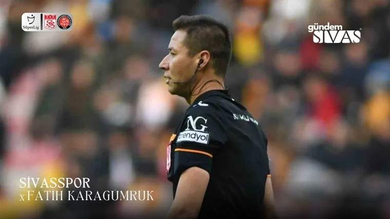 Saha kenarında kulaklık ve siyah forma takan bir hakem, bulanık tribünler önünde profilden görülüyor.